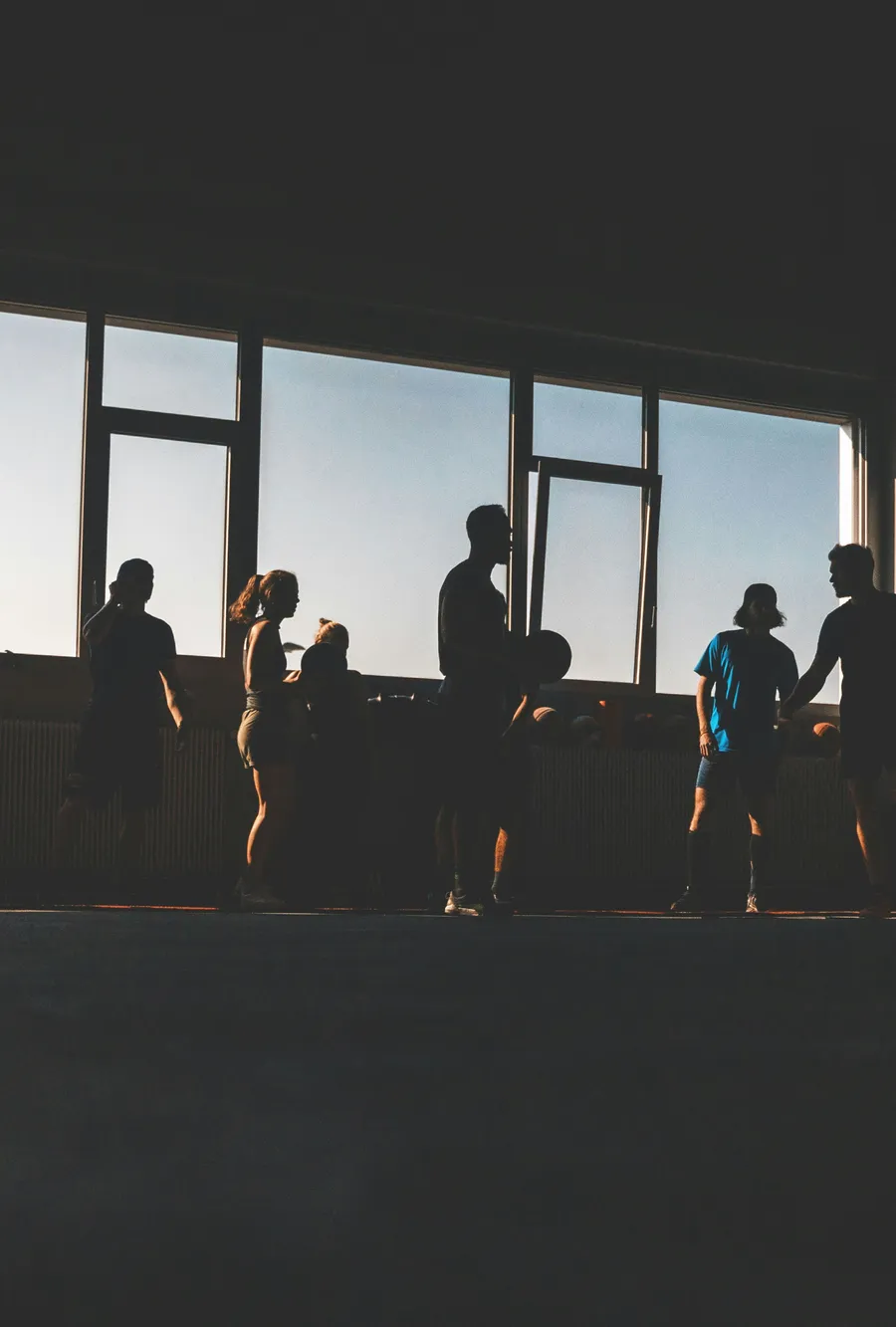 People training together in a gym, silhouetted against tall windows.