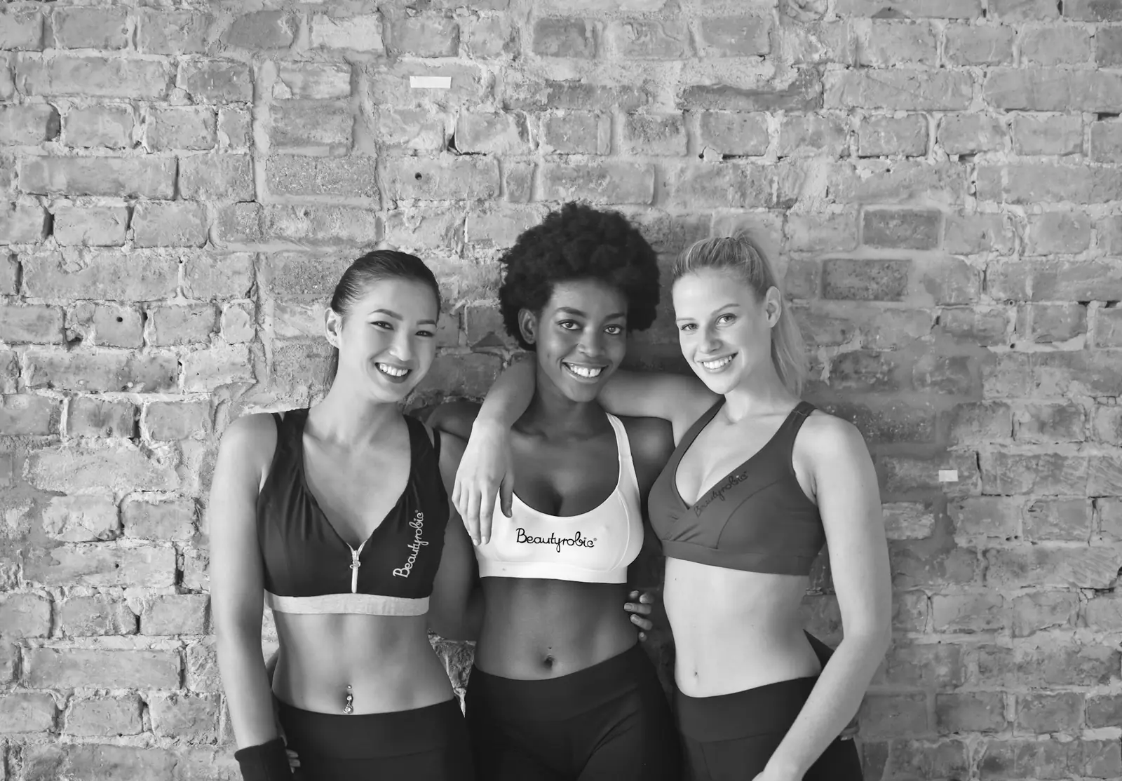 Three women in workout clothes standing arm in arm against a brick wall, in black and white.
